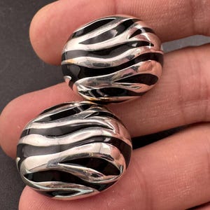 May include: A pair of round earrings with a silver finish and a black enamel wave pattern. The earrings are held in a hand, highlighting the polished surface and detailed design. The design features alternating silver and black curved lines.