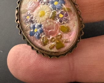 May include: Vintage oval brooch with a floral design. The brooch has a pink background with a bouquet of white, blue, and purple flowers. The metal frame has a decorative, scalloped edge.