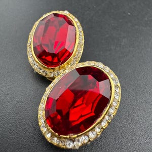 May include: A pair of vintage clip-on earrings. Each earring has a large, oval, faceted, ruby-red stone set in a gold-tone metal frame. The frame is encrusted with small, clear rhinestones. The earrings are displayed on a dark surface.