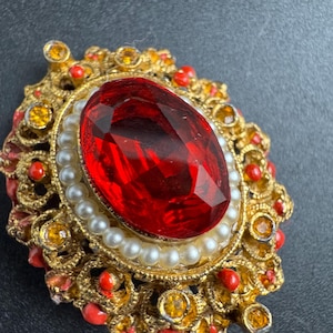 Peut inclure: Broche vintage dorée avec une grosse pierre rouge ovale et facettée au centre. La pierre est entourée d'un anneau de petites perles blanches. La broche présente des détails ornés avec des accents rouges et jaunes.