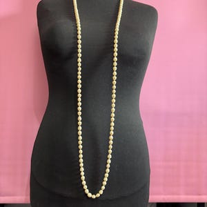 May include: A long, cream-coloured pearl necklace displayed on a black mannequin. The pearls are evenly spaced and have a subtle lustre. The background is a solid pink colour.
