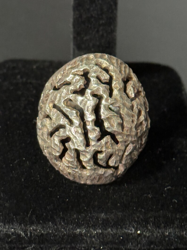 May include: A silver ring with a textured, abstract design. The ring is round and has a raised, intricate pattern.