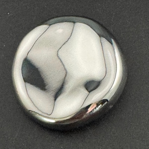 May include: A circular, silver-toned object with a highly reflective surface. The object has a smooth, polished appearance with dark, irregular patterns across its surface. The design is simple and modern, with a high-gloss finish.