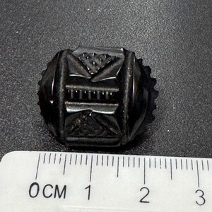 May include: A black glass button with a geometric design. The button is approximately 1 cm in diameter.