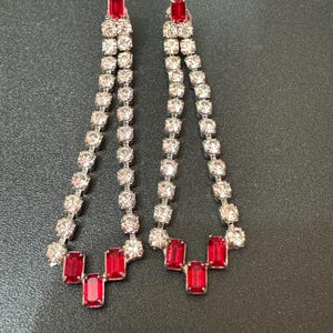 May include: A pair of long, dangling earrings with a silver chain and red rectangular stones. The earrings have a cascading design with multiple rows of stones.