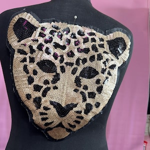 May include: A large, sequined leopard head patch in tan, black, and purple. The patch is attached to a black mannequin. The leopard's face is detailed with black sequins for the eyes, nose, and spots, and tan sequins for the rest of the face.