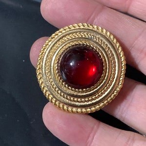 May include: A gold-toned brooch with a red cabochon stone set in a circular design. The brooch has a rope-like border.