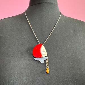 May include: A gold chain necklace with a red and white sailboat pendant. The sailboat has a small anchor on the sail and a gold chain with a small amber-colored charm hanging from the bottom.