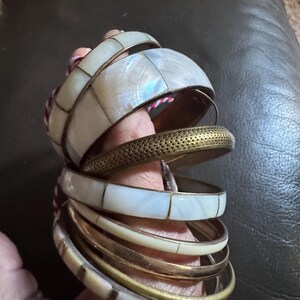 May include: A selection of bangles in various styles. Some are gold-coloured, while others have white, rectangular inlays. One bangle has a perforated pattern. The jewellery is stacked on a hand.