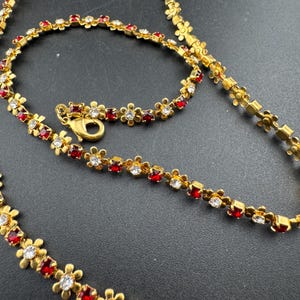 May include: Gold-coloured necklace and bracelet set with a floral pattern. Each flower is decorated with red and clear gemstones. The necklace and bracelet have a lobster clasp closure.