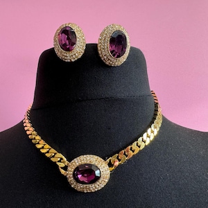 Könnte beinhalten: Eine goldene Kettenkette mit einem großen ovalen Anhänger mit einem violetten Edelstein, der von kleineren klaren Steinen umgeben ist. Passende Ohrringe mit violetten Edelsteinen und klaren Steinen.