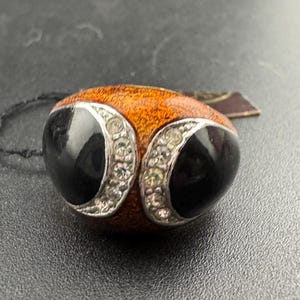 May include: A black and orange ring with two black oval stones set in a silver band. The stones are surrounded by small clear rhinestones.