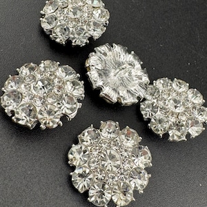 以下が含まれることがあります： 黒い表面に十字形に配置された、クリアなラインストーンがあしらわれた銀色の丸いボタン5つ