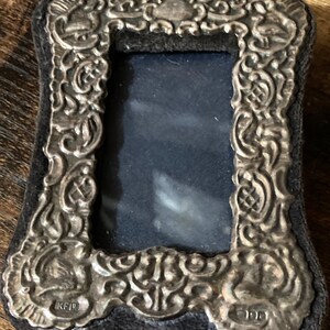 Puede incluir: Un marco de fotos plateado con un dise&ntilde;o ornamentado y un respaldo de terciopelo negro. El marco tiene una abertura rectangular para una foto y est&aacute; marcado con las letras "KFP" y "100".