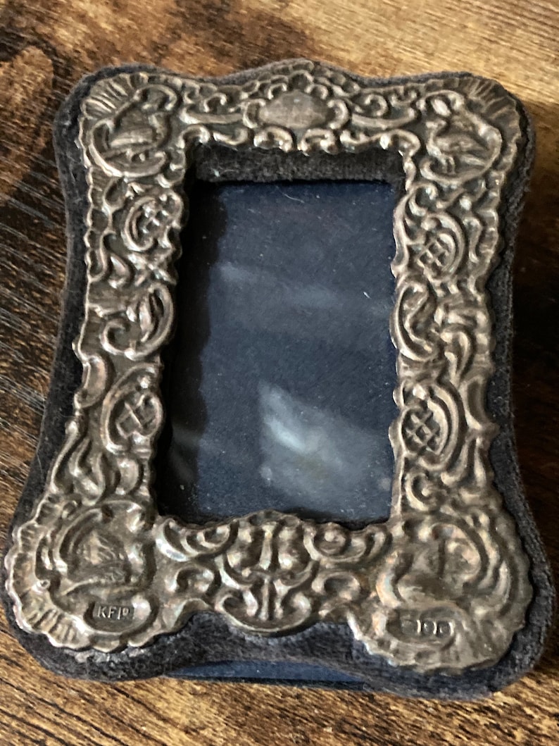 Puede incluir: Un marco de fotos plateado con un dise&ntilde;o ornamentado y un respaldo de terciopelo negro. El marco tiene una abertura rectangular para una foto y est&aacute; marcado con las letras "KFP" y "100".