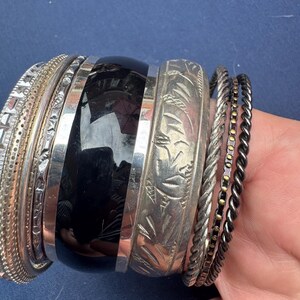 May include: A collection of stacked bracelets including a wide black resin bangle, a silver-toned bangle with etched leaf patterns, a textured silver-toned band, a twisted silver-toned rope bracelet, and a thin dark metal band with gold-toned dots.