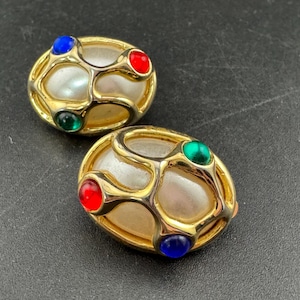 May include: A pair of gold-coloured oval clip-on earrings. Each earring has a central pearl-like feature, framed by a gold filigree pattern. The earrings are decorated with red, green, and blue gemstones, adding colour to the design.