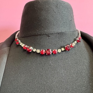 May include: A silver necklace featuring alternating red and clear gemstones. The necklace is displayed on a black mannequin head against a pink backdrop. The red stones are square-cut, and the clear stones are smaller and round.