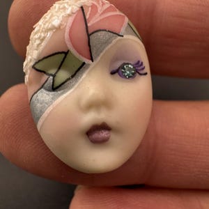 May include: A hand-painted porcelain doll face with a stylized design. The face features a pink rose, silver accents, and a purple eye with a jewel. The lips are a muted mauve color. The doll face is oval-shaped.
