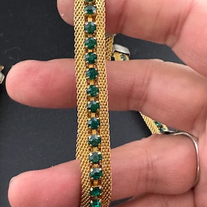 May include: Gold-toned mesh bracelet with a row of emerald green rectangular stones. The bracelet has a delicate, woven design and a secure clasp. The stones are evenly spaced and add a touch of color and sparkle to the accessory.