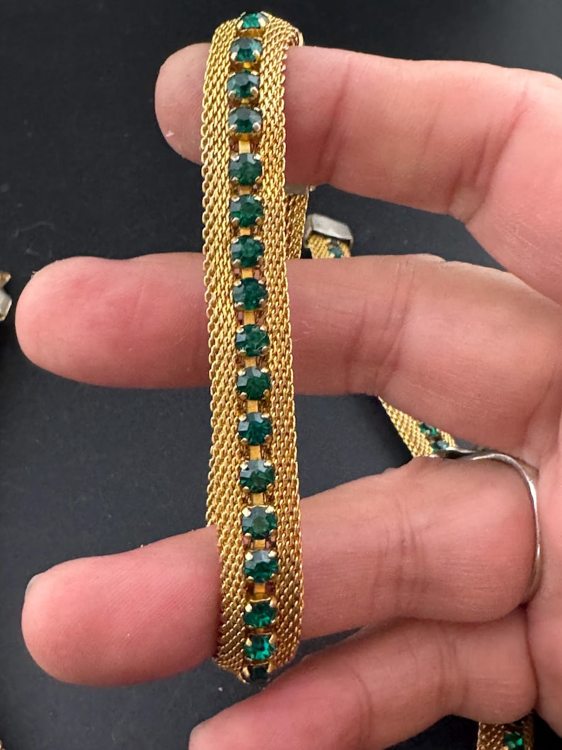 May include: Gold-toned mesh bracelet with a row of emerald green rectangular stones. The bracelet has a delicate, woven design and a secure clasp. The stones are evenly spaced and add a touch of color and sparkle to the accessory.