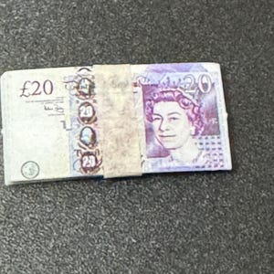 May include: A stack of twenty pound banknotes featuring a portrait of Queen Elizabeth II. The banknotes are purple, blue, and white.