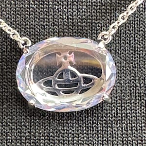 May include: Silver-toned necklace featuring a large, oval-shaped pendant with a clear, faceted crystal. The pendant has a silver-toned emblem in the center. The chain is also silver-toned. The necklace is displayed on a dark gray textured surface.