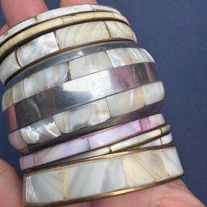 May include: A stack of five bangle bracelets featuring inlaid iridescent mother-of-pearl segments. The bangles are constructed with alternating bands of silver-toned and gold-toned metal, showcasing a variety of white, cream, and pinkish hues.