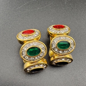 May include: A pair of gold-coloured earrings with oval-shaped red and green gemstones. Each earring features a ring of clear crystals around the gemstone and on the base.
