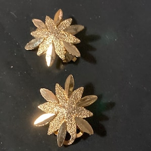 May include: A pair of gold-tone clip-on earrings with a flower design. The earrings have a textured surface and are made of metal.