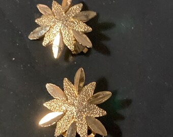 May include: A pair of gold-tone clip-on earrings with a flower design. The earrings have a textured surface and are made of metal.