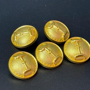 May include: Five gold-coloured buttons featuring a horse bit design. The buttons are round with a textured surface and a raised bit detail in the centre. The buttons are approximately 2.5 cm in diameter.