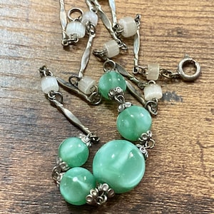 Pu&ograve; includere: Una collana a catena d'argento con perline di vetro verde e bianco. La collana ha una chiusura a moschettone.