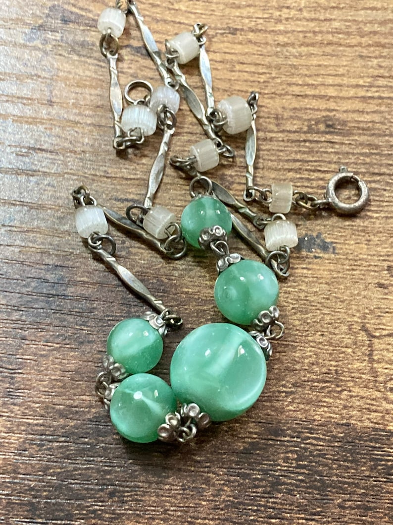 Pu&ograve; includere: Una collana a catena d'argento con perline di vetro verde e bianco. La collana ha una chiusura a moschettone.