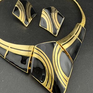 May include: A vintage jewelry set featuring a gold-tone necklace and matching earrings. The set has a geometric design with black enamel and gold accents. The necklace is a collar style, and the earrings are square-shaped.
