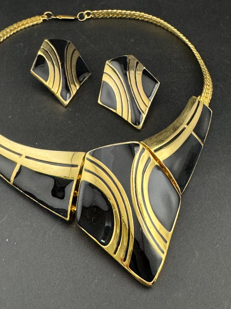 May include: A vintage jewelry set featuring a gold-tone necklace and matching earrings. The set has a geometric design with black enamel and gold accents. The necklace is a collar style, and the earrings are square-shaped.