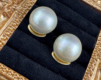 May include: A pair of large, round pearl earrings with a lustrous, iridescent sheen. The pearls are set on small, gold-colored bases, displayed against a black velvet background within a decorative gold frame.