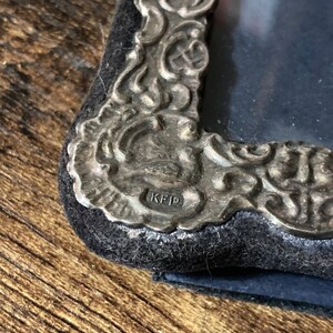 Puede incluir: Primer plano de un marco de fotos plateado con un dise&ntilde;o ornamentado. El marco tiene un respaldo de terciopelo negro y la inscripci&oacute;n "KFD" es visible en la parte inferior.
