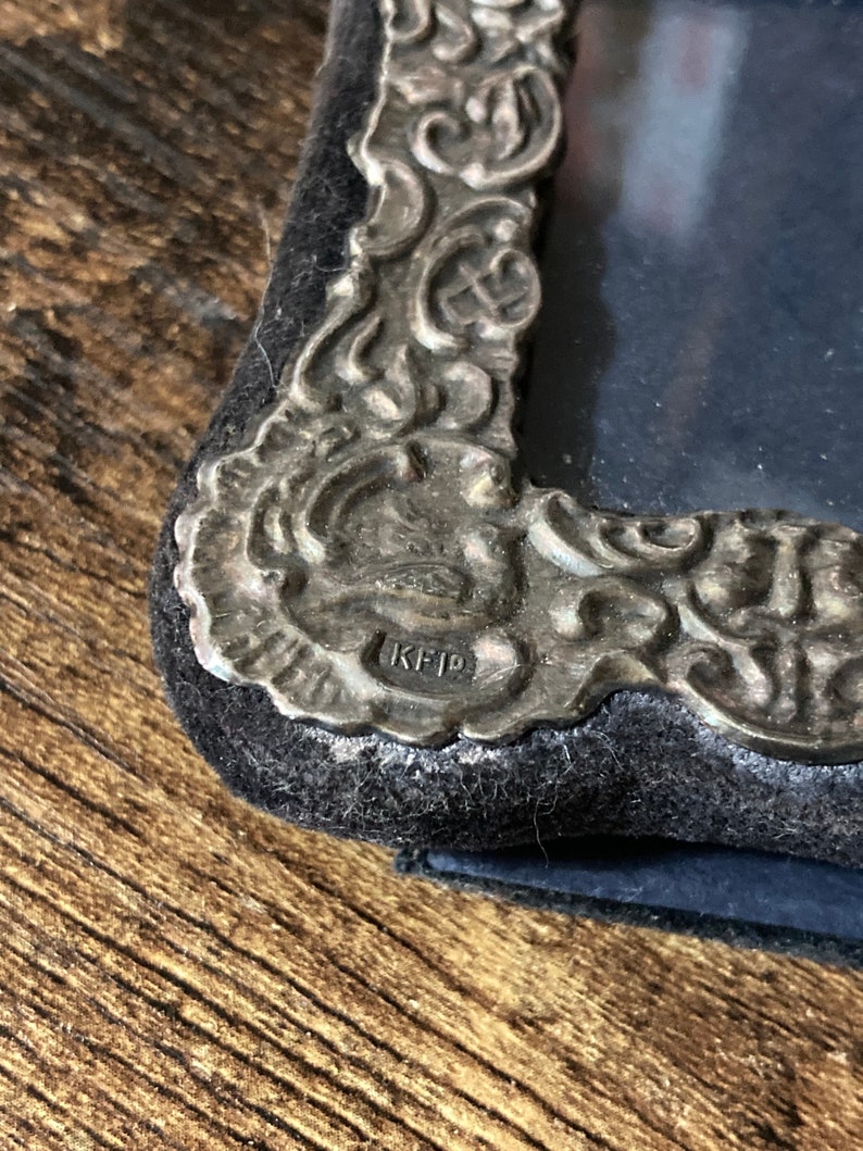 Puede incluir: Primer plano de un marco de fotos plateado con un dise&ntilde;o ornamentado. El marco tiene un respaldo de terciopelo negro y la inscripci&oacute;n "KFD" es visible en la parte inferior.