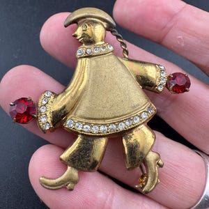 May include: Vintage gold-tone metal brooch in the shape of a figure with a hat and coat. The brooch is embellished with clear rhinestones and two large red gemstones. The figure is holding a red gemstone in each hand.