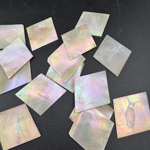 May include: A collection of iridescent square mother-of-pearl tiles. The tiles have a pearlescent sheen with hints of pink, green, and gold. The squares are scattered on a dark surface, showcasing their natural beauty and potential for craft projects.
