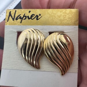 Könnte beinhalten: Ein Paar goldfarbene Clip-Ohrringe mit einem strukturierten, blattartigen Design. Die Ohrringe befinden sich auf einer weißen Karte mit dem Markennamen "Napier" in Schwarz. Die Ohrringe haben eine polierte Oberfläche.