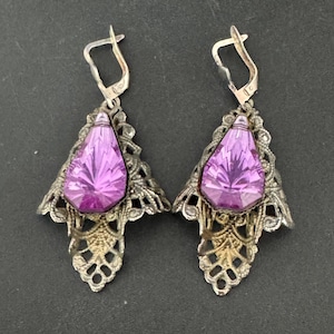 May include: A pair of vintage earrings featuring large, faceted, teardrop-shaped purple gemstones. The gemstones are set in ornate, filigree-style silver-toned metalwork. The earrings have a lever-back closure.