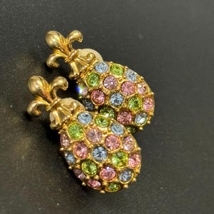 May include: A pair of gold-tone earrings with a fleur-de-lis design. The earrings are covered in multi-coloured rhinestones.