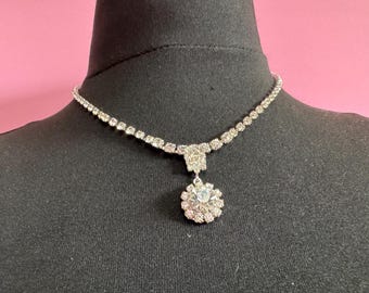 May include: A silver-toned necklace with a sparkling design. The necklace features a delicate chain of small, round crystals. A square-shaped crystal cluster connects the chain to a larger, round pendant with a floral design, also made of crystals.