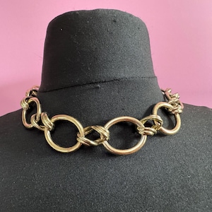 May include: A gold-toned chain necklace with a series of interlocking oval links and twisted accents. The necklace is displayed on a black mannequin against a pink background. The jewelry has a bold, chunky design.