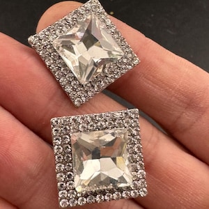 May include: Two square-shaped, silver-toned earrings. Each earring features a large, clear, square-cut crystal center, surrounded by a border of smaller, clear crystals. The earrings are set against a dark background.