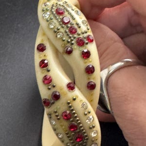 May include: Ivory-colored, intertwined bracelet adorned with red and clear gemstones. The bracelet features a detailed pattern of small, round red stones and clear crystals, set against a gold-toned metal. A vintage-style accessory.