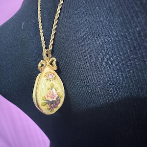 May include: A gold chain necklace with a pendant shaped like an egg. The egg is decorated with a floral design and a small pink rhinestone.