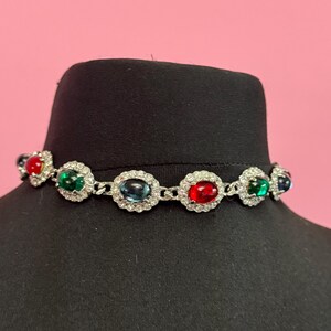 May include: A silver choker necklace with a chain link design and five oval-shaped gemstones. The gemstones are red, green, blue, and green. The necklace is on a black mannequin.
